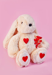 Romantic plush bunny sits on a pink background with bouquet of flowers and red hearts on paws, valentine's day card concept, love relationship