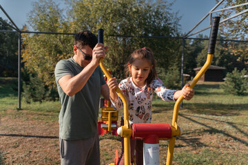Obraz premium A father assists his daughter while they use an outdoor exercise machine together, enjoying a fun fitness moment