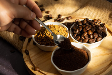 Female hand holding a spoon scooping ground coffee from a small bowl next to different types of coffee beans and instant coffee receiving the morning sun. Coffee preparation