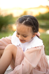 A gentle summer picnic by the water. Portrait of a blue-eyed girl with long hair in a light dress on the grass and on a beige blanket with flower and fruit. A gentle rest. Warm evening