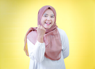 A smiling asian woman wearing a hijab celebrates with raised hands against a bright yellow background