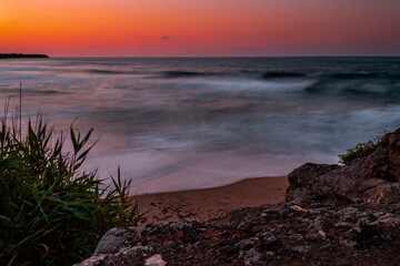 Long exposure creating soft waves gently lapping the shoreline during a vibrant sunset. Grass, rocks bushes and sand on the beach. Colorful sky