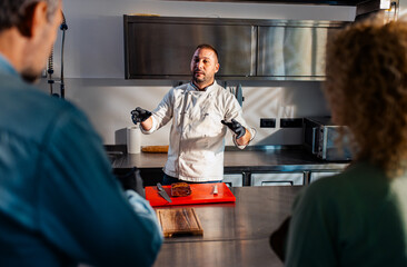 Chef in the kitchen holding a cooking class to a couple, showing how to cut bacon.