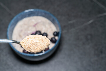Spoon with a healthy dietary supplement, like protein and maca powder, is given into a bowl with muesli. Bowl with muesli is blurry in the background, containing cereals, banana, blueberry and others.