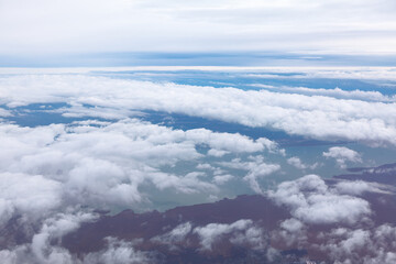 Aerial view of clouds stretching over landscape, with glimpses of land and water below. Sky displays soft gradients of blue and white creating a serene and atmospheric scene from high altitude