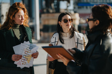 Three businesswomen engage in an outdoor discussion about project plans and data analysis charts. They share documents and collaborate in a lively, professional atmosphere in a sunny urban setting.