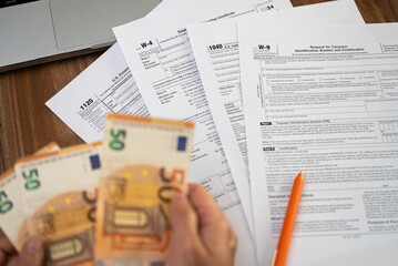 Obraz premium Close-up of hands holding euro bills over various tax forms including W-4, 1040, W-9, and 1120. Tax season, financial accounting, and tax refund concept 