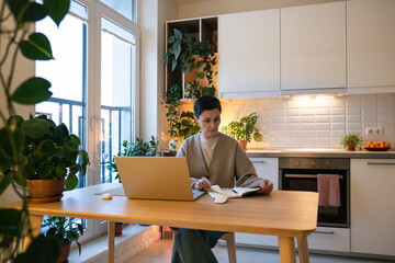 Fototapeta premium Mature woman reading notebook entries and working on laptop in cozy kitchen. Female focused on studies, preparing materials for lessons and research, adult student balancing work and learning at home