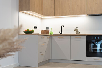 Small white kitchen with stylish, wooden cabinets, oven and sink with faucet. Appliances on the counter.  Bright interior of room in new modern apartment.