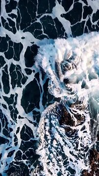 Top-down capture of waves breaking on a rocky shore, water patterns resembling marble. Cool blues and whites with texture, conveying natural power.
