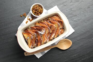 Freshly baked bread pudding in baking dish, raisins and wooden spoon on dark textured table, top view