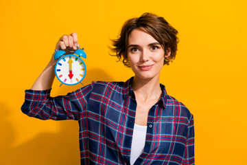 Photo of positive cheerful adorable woman with bob hairdo dressed plaid shirt holding clock in arm isolated on yellow color background