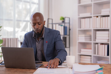 Naklejka premium Focused african american manager analyzing financial report using laptop and reading documents while sitting at desk in modern office, busy entrepreneur working with computer and paperwork