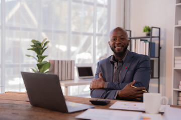 Portrait of a smiling african american businessman sitting at his desk with crossed arms in a luminous office, with laptop, calculator, documents and a cup of coffee