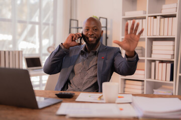 Fototapeta premium Cheerful African American businessman engaging in a phone call while waving to someone, sitting at his desk in a bright, modern office filled with natural light