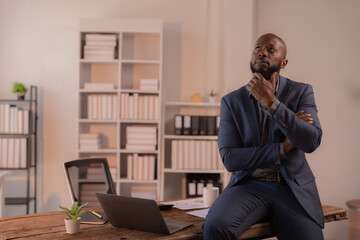 Thoughtful african american businessman in suit sitting on desk in modern office looking up and thinking about future business strategies, planning new projects, and pondering over ideas