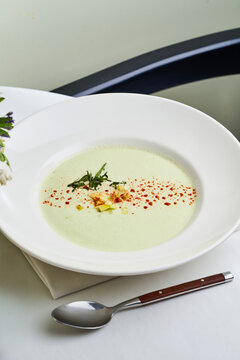 A vibrant green soup is elegantly presented in a white bowl, garnished with herbs, spices, and bits of ingredients, showcasing a fresh culinary creation at lunch
