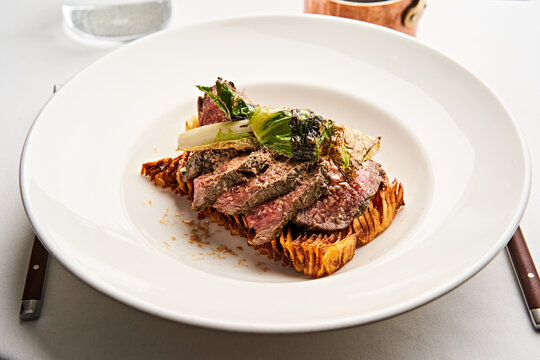 Succulent grilled steak slices rest on a toasted bread base, complemented by charred greens and garnishes, all arranged elegantly on a white plate