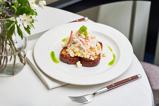 A delicious seafood salad is presented on toasted bread, topped with vibrant garnishes. The dish is surrounded by a dining setting featuring fresh flowers, creating a refined atmosphere