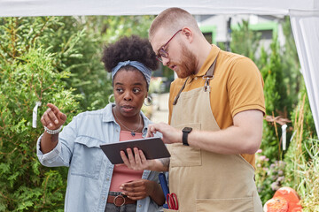 Medium shot of African American female landscape designer pointing at exact coniferous shrub choosing plants for garden design, while talking to male horticulturalist using digital tablet outdoors