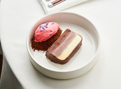 A delicate dessert is presented on a white plate, featuring a layered chocolate cake beside a scoop of pink sorbet, garnished with lavender. Enjoying a refined dining experience