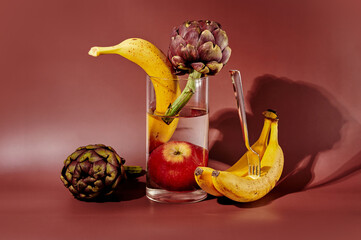 Surreal still life composition with bananas, artichokes, and an apple in a glass of water, alongside a fork stabbed into a banana. Conceptual food art against a deep red background