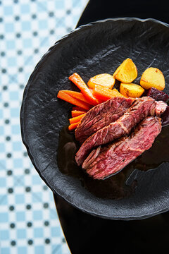 A gourmet steak is beautifully plated alongside roasted carrots and potatoes. The dish is presented on a chic black plate, set against a fashionable backdrop