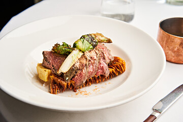 Plated beef dish showcases tender herb-crusted meat atop roasted vegetables, garnished with fresh greens, served in an elegant dining environment