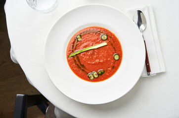 A vibrant bowl of tomato soup is artfully presented with slices of cucumber and a sprig of asparagus on a white tablecloth, showcasing culinary creativity in an elegant setting