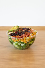 Side view of a Southwest salad with black beans, roasted corn, tortilla strips, avocado, and pepitas in a clear plastic bowl on a wooden countertop