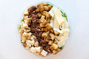 Classic Caesar salad with grilled chicken, bacon, croutons, and shaved parmesan in a clear plastic bowl