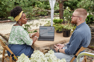 Side view of professional female landscape architect of Black ethnicity sitting at garden table with laptop displaying calendar page for planning, talking to male client about design project outdoors
