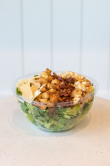 Side view of a Caesar salad with grilled chicken, croutons, bacon, and shaved parmesan in a clear plastic bowl on a white table