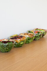 Assorted fresh salads in clear plastic bowls lined up on a wooden countertop with a minimal white background
