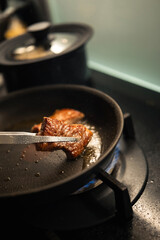 Seared Wagyu beef cube held with tongs over a sizzling black nonstick frying pan on a gas stove, capturing the rich marbling and golden crust