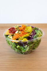 Side view of a fresh garden salad with broccoli, shredded carrots, banana peppers, cucumbers, and red cabbage in a clear plastic bowl on a wooden countertop