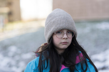 Sad almost crying teenage girl in glasses, hat and winter jacket outdoors. Concept of hard childhood