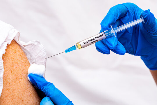 The picture shows a doctor holding a syringe with the typhoid fever vaccine in her hand, wearing blue gloves, and in the background is an old woman waiting to be vaccinated with this preparation.