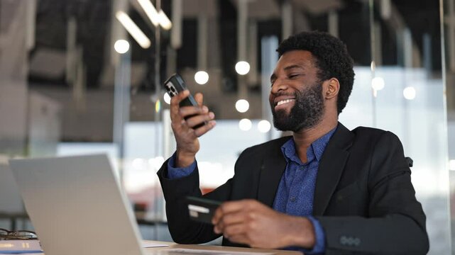 Cheerful young African businessman making online payment on smart phone. Modern office environment. Black male entrepreneur shopping on internet.