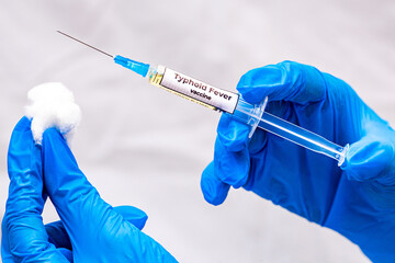 The picture shows a doctor holding a syringe with the typhoid fever vaccine in her hand, wearing...