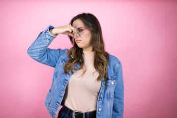 Young beautiful woman wearing denim jacket over isolated pink background smelling something stinky and disgusting, intolerable smell, holding breath with fingers on nose