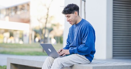 Young hispanic student working on laptop sitting on bench in university campus