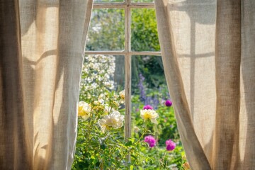 Textured beige linen curtains on a vintage window, overlooking a lush garden filled with blooming flowers.