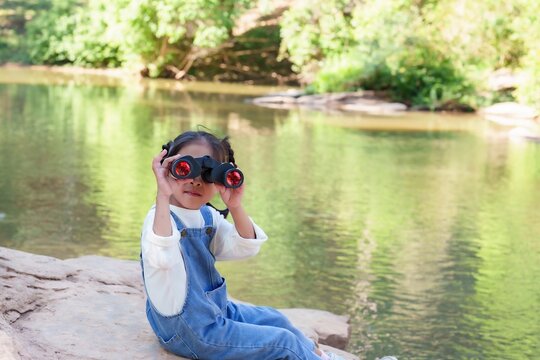 Cute Asian girl using binoculars watch birds. Enthusiastic young explorer holds binoculars while sitting on rocky surface near calm river, observing nature and enjoying the beauty of wildlife.