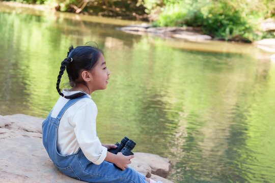 Cute Asian girl using binoculars watch birds. Sitting near peaceful river, young adventurer enjoys outdoor exploration surrounded by nature, feeling joy and curiosity in a relaxing environment.