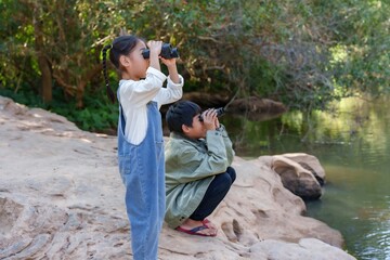 Two cute Asian brothers and sisters use telescope watch birds. Enthusiastic young adventurers observe wildlife near a flowing river, experiencing nature’s wonders in a scenic forest setting.
