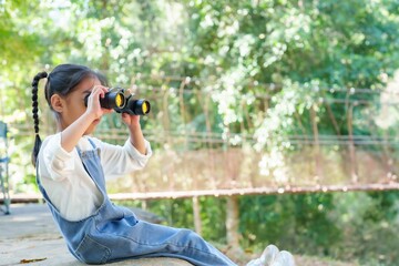 Cute Asian girl using binoculars watch birds. Sitting near a wooden bridge, young explorer gazes through binoculars, captivated by the beauty of wildlife in a lush green forest.