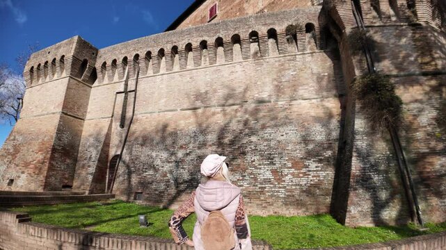 Blonde tourist walking near the Rocca Sforzesca of Dozza, medieval fortress in Emilia Romagna, is admiring the ancient walls and the blue sky in a sunny winter day