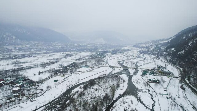 Wide angle aerial drone shot flying over the frozen Lidder river with the snow covered himalaya mountains on the side with road at the base and small villages in gulmarg kashmir