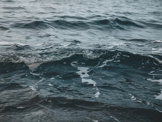  Close-up of the ocean's surface under a cloudy sky, featuring deep gray-blue waves with silvery reflections. White foam accents the dynamic water, blending shadowed depths